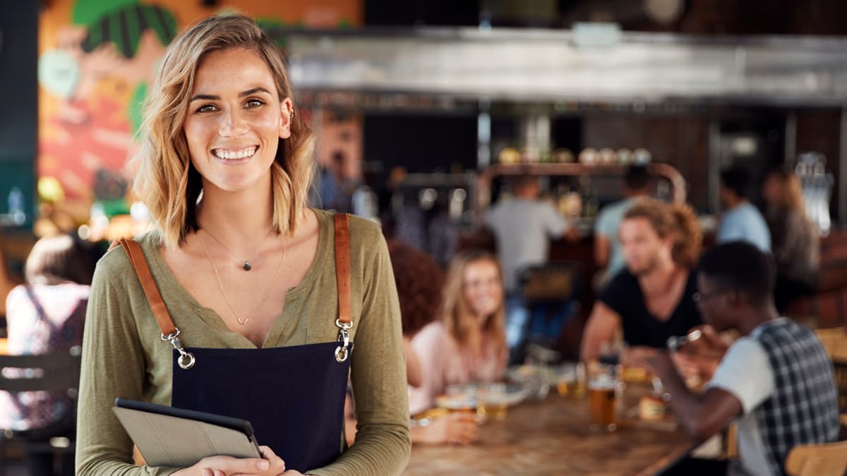 Smiling restaurant owner holding a tablet, with diners gathered behind her